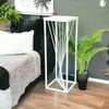 A white metal plant stand with a twisted design is placed next to a black sofa with a white pillow and a beige throw blanket. A green potted plant sits next to the stand. Anteprima dell’immagine prodotto