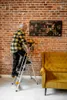 An elderly man is standing on a stepladder, reaching towards a painting on a brick wall. A yellow sofa is visible to the right. Anteprima dell’immagine prodotto