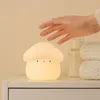 A hand reaching towards a mushroom-shaped night light on a shelf, with a baby bottle and blankets in the background. Podgląd zdjęcia produktu