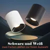 Two modern cylinder-shaped LED lights, one white and one black, are displayed at an angle against a dark, abstract background resembling rolling hills or dunes. Produktbild-Vorschau