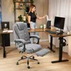 A woman is standing at a desk in a home office, holding a piece of paper. A gray ergonomic office chair with a footrest is in the foreground. Produktbild-Vorschau