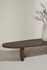 A close-up, studio shot of a dark brown, oval-shaped coffee table with a single leg. A vase with dried flowers is placed on the right side of the table. Produktbild-Vorschau