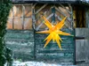 A yellow LED star lantern hangs in front of a weathered wooden shed in a snowy outdoor setting. Produktbild-Vorschau