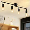 A black track light fixture with four exposed bulbs is mounted on a white ceiling above a wooden shelf with books and a small vase. Voir les images du produit