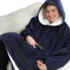 A woman is sitting on a couch, wearing a navy blue hoodie blanket and holding a remote control. Náhled obrázku produktu