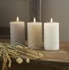 Three LED candles of varying shades of gray and white, with a textured swirl pattern, are placed on a wooden surface next to dried grass. Produktbild-Vorschau