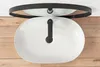 A white oval sink with a black faucet is shown from a top-down view, placed on a wooden surface. A black framed mirror is visible above the sink. Náhled obrázku produktu