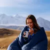 A young woman wrapped in a blue sleeping bag, holding a mug, in a mountainous landscape. Produktbild-Vorschau