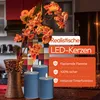 Three blue LED candles with lit flames are placed on a wooden surface next to a decorative vase with orange flowers. The background shows a kitchen setting. Voir les images du produit