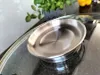 A close-up of a stainless steel lid with a built-in steam vent, resting on a black speckled surface. A potted plant and a yellow bell pepper are visible in the background. Produktbild-Vorschau