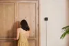 A woman in a yellow dress is opening a wooden door, with a black smart home device mounted on the wall to the right. Anteprima dell’immagine prodotto