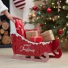 A person is placing a gift into a Christmas-themed sleigh filled with presents, with a decorated Christmas tree in the background. Produktbild-Vorschau