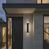 A modern house entrance at dusk, featuring a black front door and a sleek wall-mounted light fixture. Náhľad obrázku produktu