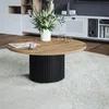 A round coffee table with a wooden top and a black fluted base, placed on a fluffy white rug in a living room setting. A vase with white flowers and a magazine are on the table. Náhľad obrázku produktu