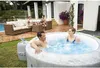 A couple relaxing in an inflatable hot tub in a backyard setting, with other people and outdoor furniture in the background. Produktbild-Vorschau