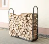 A black metal firewood rack filled with chopped wood is shown outdoors on a paved surface next to a building. Náhled obrázku produktu