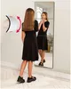 A young girl in a black dress stands in front of a mirror, looking at her reflection. An overlay graphic shows hands bending a curved white object with red arrows indicating movement. Anteprima dell’immagine prodotto