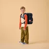 A young boy wearing a backpack and standing against a plain background. Produktbild-Vorschau