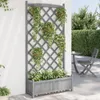 A grey wooden planter box with a trellis attached, filled with green plants, stands against a white wall. A grey garden chair and table are visible in the background. Produktbild-Vorschau