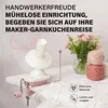 A yarn winder and balls of yarn are displayed on a white surface, with a bouquet of pink flowers in the background. Produktbild-Vorschau