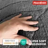 A hand is shown pressing into a soft, dark gray bath mat, highlighting its texture and plushness. Several logos and text overlays are present, indicating product features and brand. Produktbild-Vorschau