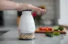 A hand pressing down on a white manual food chopper filled with nuts, with chopped vegetables in the background. Produktbild-Vorschau