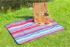 A picnic blanket laid out on grass next to a body of water, with a picnic basket and some fruit on the blanket. Podgląd zdjęcia produktu
