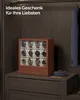 A wooden watch box with 12 watches displayed on a dresser, next to a gift-wrapped box, with a suit hanging in the background. Produktbild-Vorschau