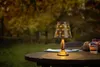 A solar-powered table lamp sits on a wooden table outdoors, with a blurred background of trees and foliage. Anteprima dell’immagine prodotto