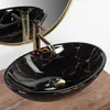 A black marble sink with gold veins and a gold faucet is shown on a wooden surface, with a gold-rimmed mirror in the background. Produktbild-Vorschau