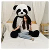 A young woman is sitting on a bed next to a very large stuffed panda. Produktbild-Vorschau