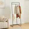 A modern black metal coat rack with two sweaters and a coat hanging on it, placed in a living room next to a sofa. Produktbild-Vorschau