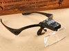 A black head-mounted magnifier with a detached lens lies on a textured surface, with a wooden background behind it. Náhled obrázku produktu