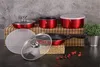 A set of red cookware, including pots and a frying pan, is arranged on a bamboo mat and a dark surface. A potted plant is visible in the background. Podgląd zdjęcia produktu
