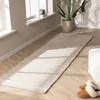 A long, narrow rug is laid out on a wooden floor in front of a window. A potted plant is visible near the window, and a pair of slippers are placed on the rug. Podgląd zdjęcia produktu