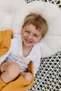 A young child is lying on a white cushion, wrapped in a mustard yellow blanket. Produktbild-Vorschau