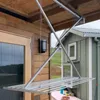 A retractable clothes drying rack is mounted on the ceiling of a covered porch, with a wooden wall and door on the left and a green building on the right. Podgląd zdjęcia produktu