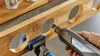 A Dremel rotary tool is being used to cut a circular hole in a wooden beam, with another hole already cut and a clamp holding the beam in place. Produktbild-Vorschau