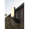 A modern outdoor wall light mounted on a wooden fence, with foliage in the foreground. Produktbild-Vorschau