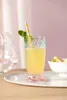 A tall glass of yellow liquid with a straw and mint garnish, placed on a white tray with a bowl of fruit in the background. Produktbild-Vorschau