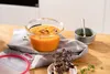 A glass food storage container filled with orange soup is placed on a gray napkin on a wooden table. A small bowl of olive oil and a sprig of microgreens are also visible. Náhľad obrázku produktu