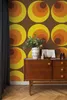 A retro-style wallpaper with concentric circles in yellow, orange, and brown is shown behind a wooden sideboard. The sideboard has a vase with greenery, framed pictures, and binoculars on top. Produktbild-Vorschau