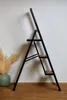 A black folding step stool with wooden steps is shown against a white wall and on a wooden floor. Anteprima dell’immagine prodotto