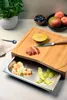 A bamboo cutting board with a pull-out stainless steel tray filled with fruit peels and chopped fruit, with whole fruits and blueberries in bowls in the background. Anteprima dell’immagine prodotto