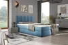 A bedroom scene featuring a blue upholstered boxspring bed with a mattress and topper, a bedside table with a lamp, and artwork on the wall. Produktbild-Vorschau
