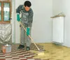 A man in overalls and green gloves is applying a yellow liquid to a checkered floor with a mop. There is a paint can on the floor and a radiator on the wall. Produktbild-Vorschau