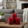 Three red LED candles are arranged on a wooden table in a living room setting, with a sofa and fireplace in the background. Produktbild-Vorschau