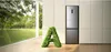 A modern stainless steel refrigerator stands in a bright, airy room with large windows overlooking a lush green landscape. A stylized 'A' shape made of green foliage is in the foreground. Produktbild-Vorschau