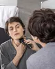 A man is shaving his neck in front of a mirror in a bathroom. Produktbild-Vorschau