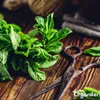 A bunch of fresh mint leaves are arranged on a wooden surface next to a pair of old scissors. In the background, a spool of twine and some folded fabric are visible. Náhľad obrázku produktu